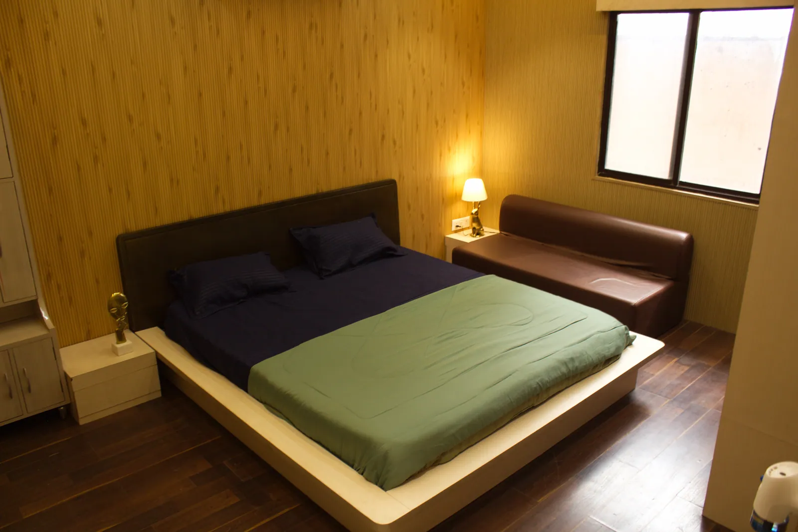 A bedroom at Raj Organic Farm with a low double bed, a green bedspread, a brown sofa and a bedside lamp